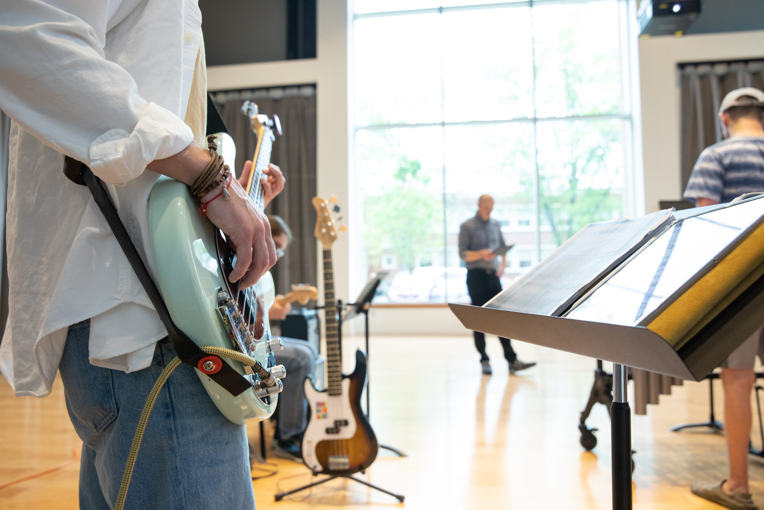 A person plays an electric bass guitar at a Haun Rehearsal while others are present in a bright room with large windows.