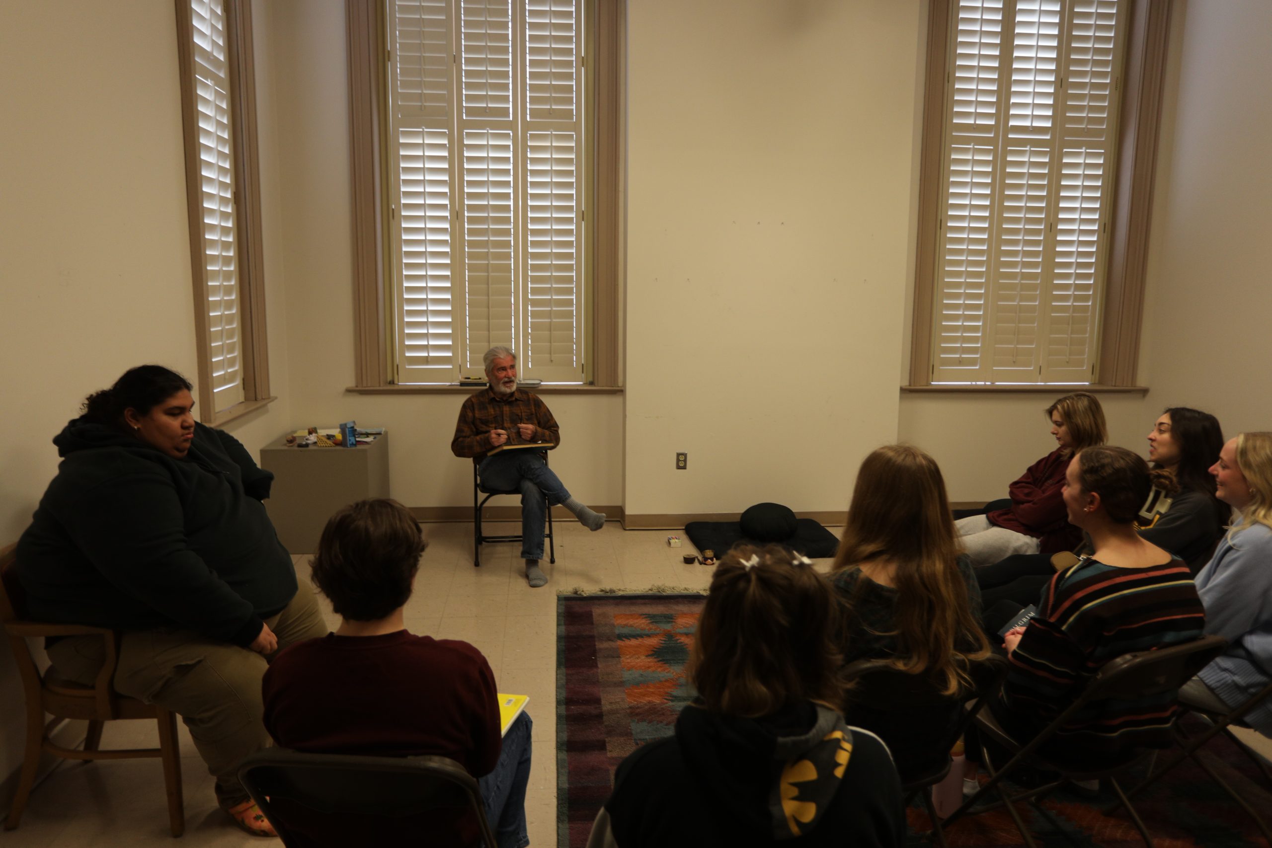 A group of people sits in chairs arranged in a circle in a room with tall, historic windows, while one person speaks to the group from a chair at the front.