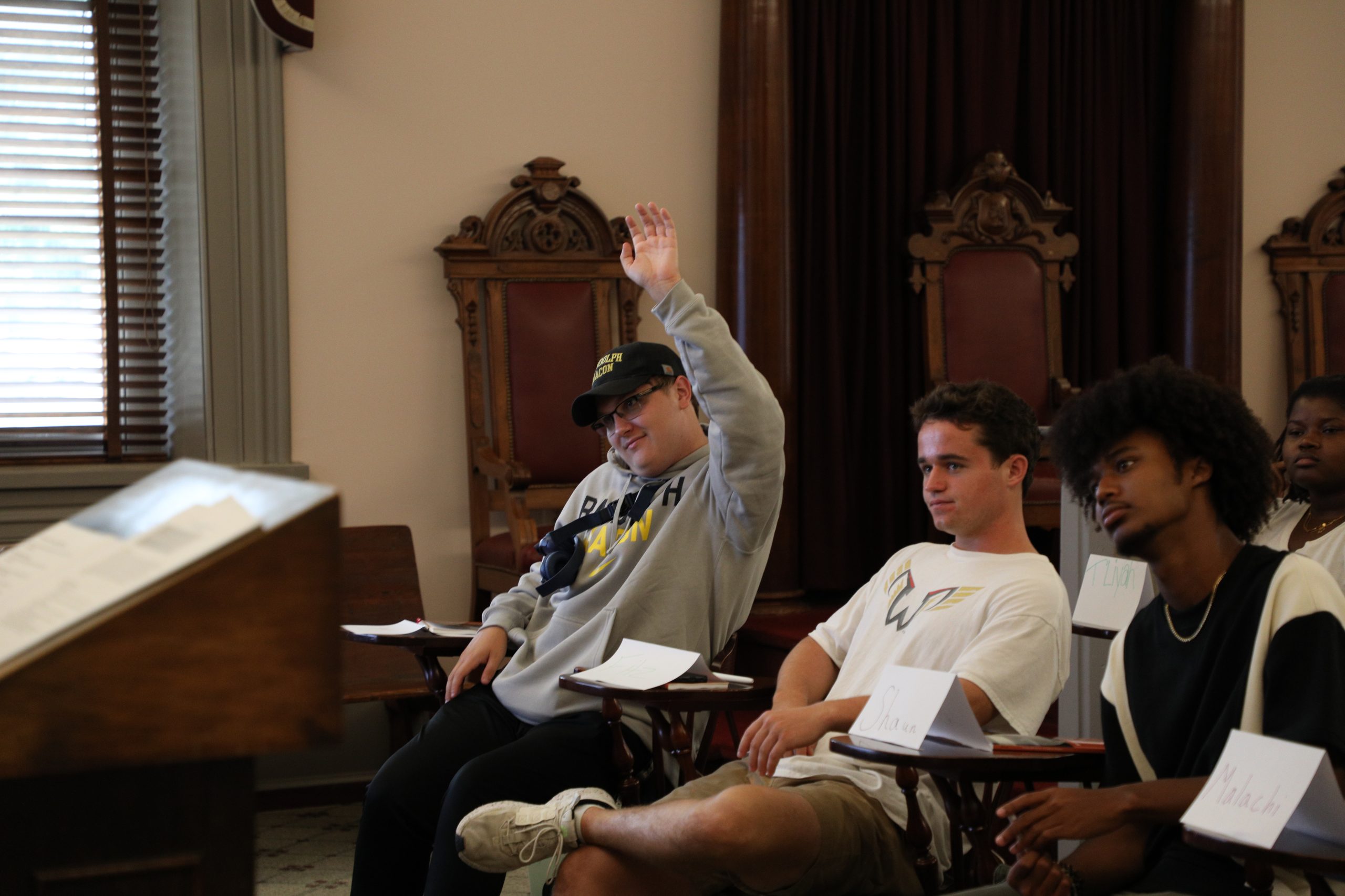 In a historic classroom on campus, a student raises his hand while sitting at a desk. Two other students sit nearby, one looking towards the front, the other gazing sideways.