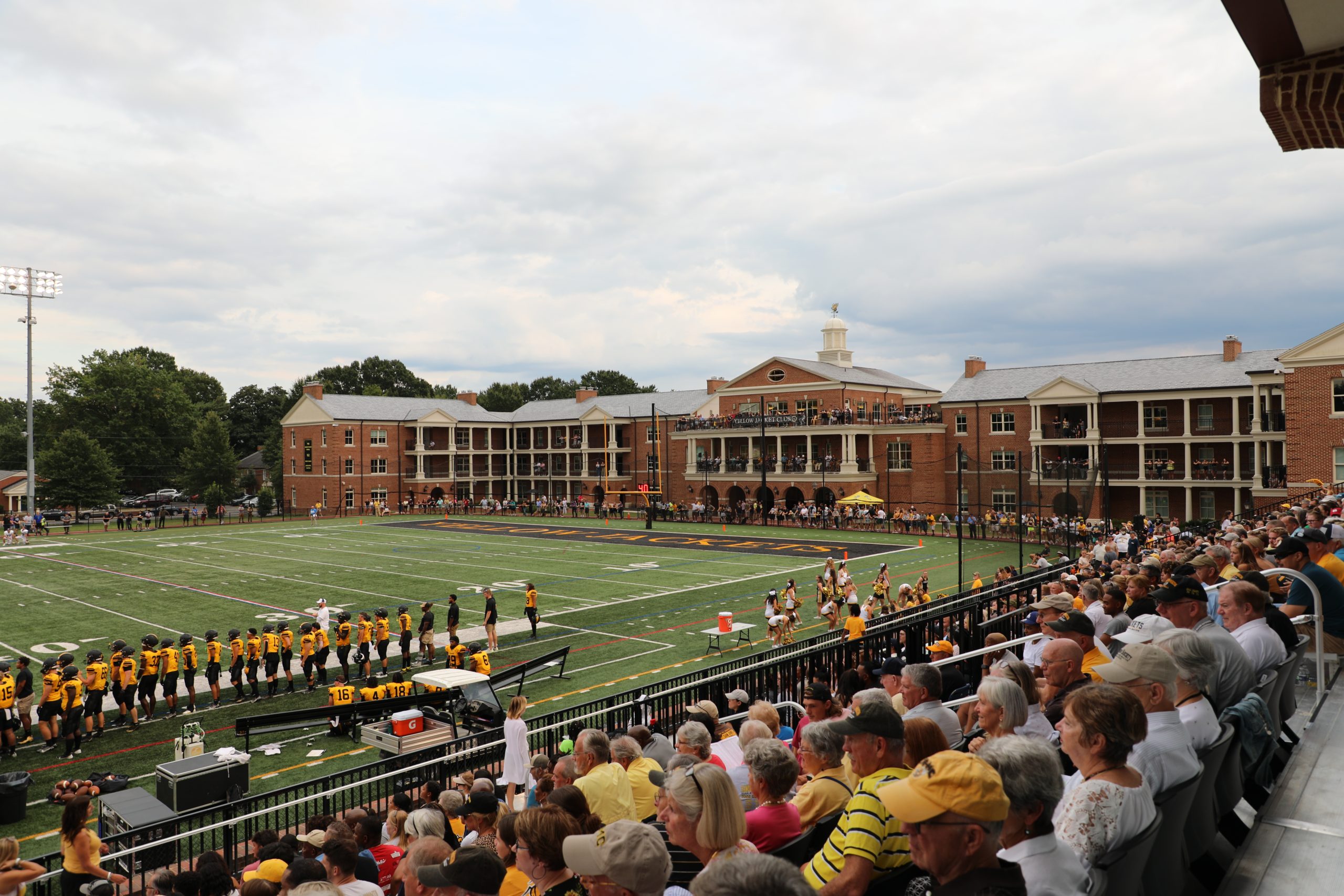 Spectators watch a football game in progress at an outdoor stadium with RMC players in yellow jacket player uniforms on the field