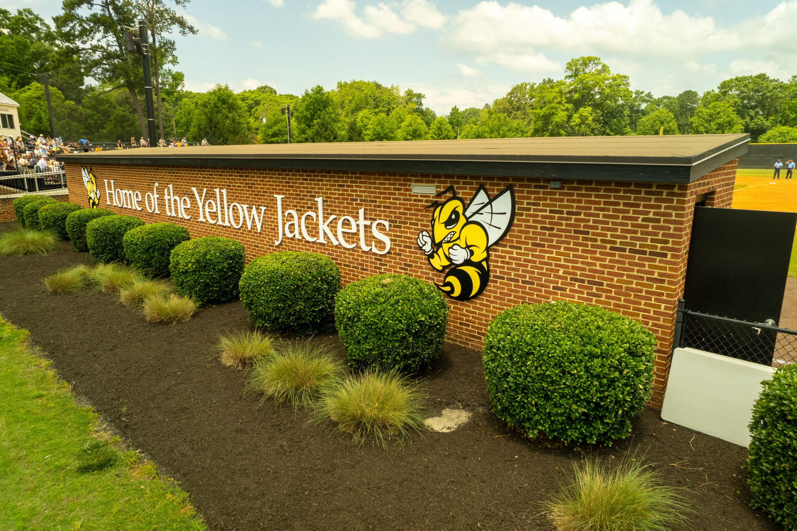 Red brick wall with the text "Home of the Yellow Jackets" and a yellow jacket mascot graphic, bordered by greenery. In the background, a baseball field in the park and spectators are visible.