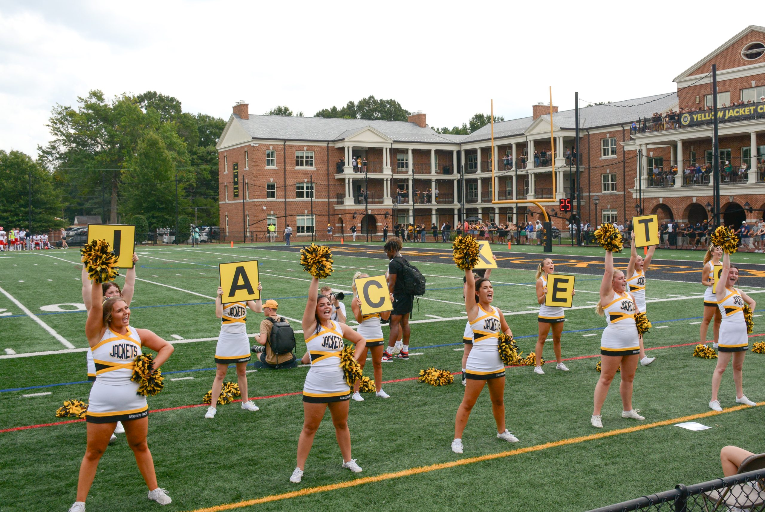RMC Cheerleaders in yellow and white uniforms hold up signs spelling "JACKETS" on a football field, with Day field in the background.
