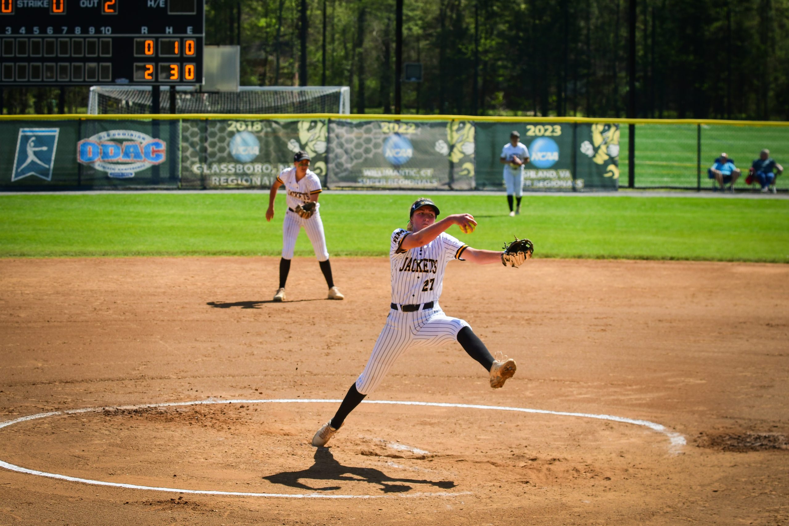 An RMC softball pitcher in mid-throw on a sunny day, with other players scattered across the field and an electronic scoreboard in the background, capturing the spirit of sports.