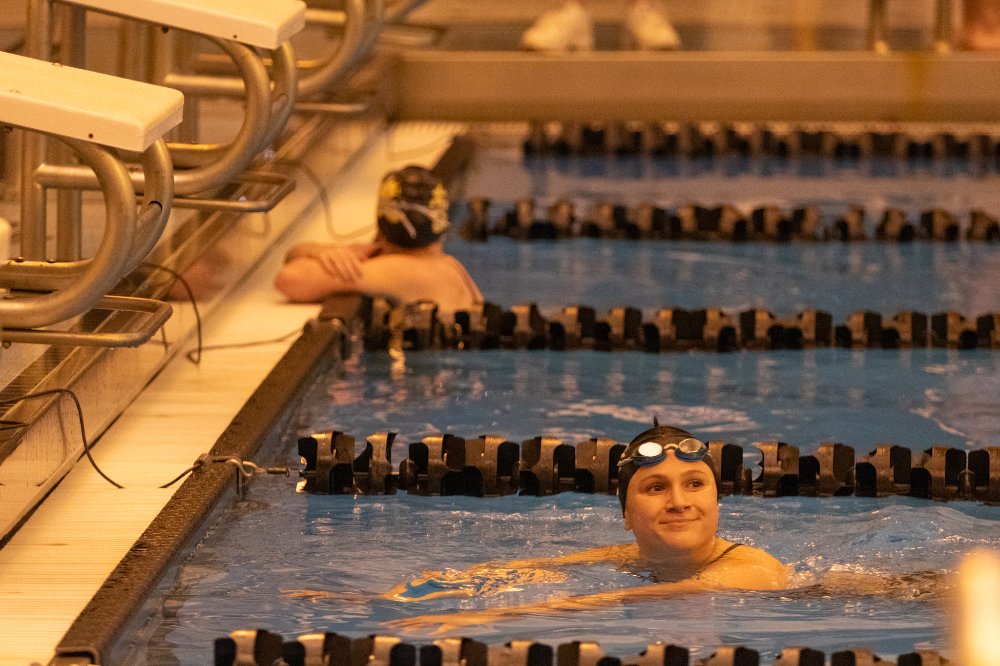 Two swimmers in a pool, one in the center foreground smiling with goggles on the head, and another in the background wearing a swimming cap and goggles, resting at the pool's edge. The scene captures a moment of pure recreation and joy.