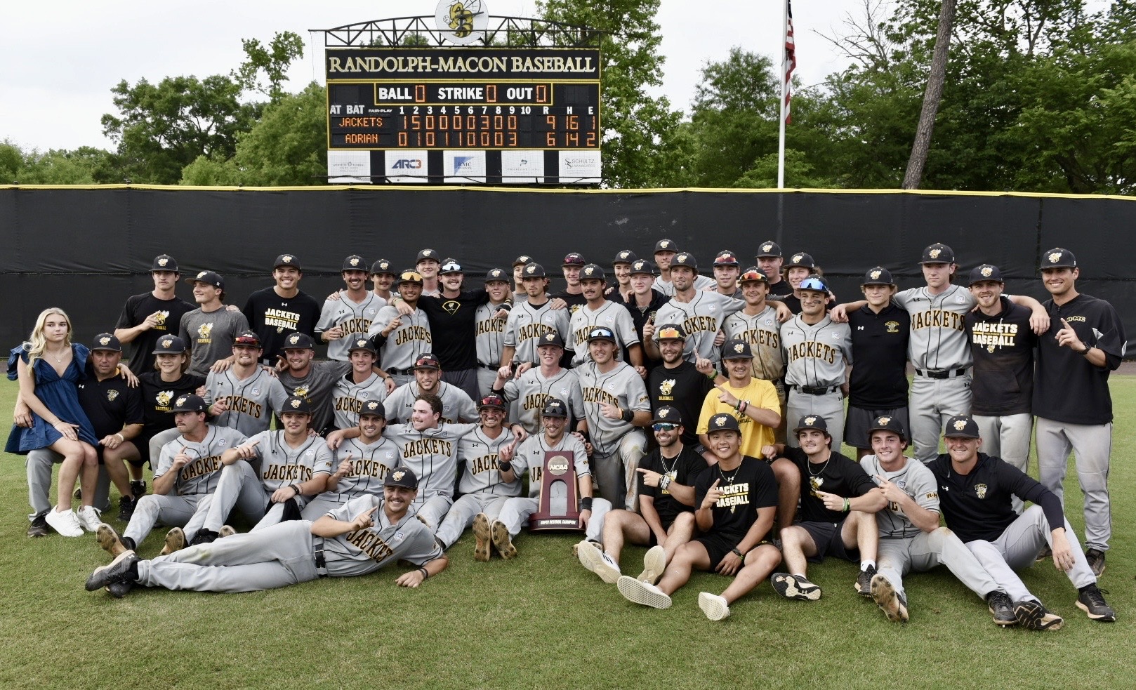 A large group of RMC baseball players and staff from Randolph-Macon College pose together on a park field, holding a trophy. The scoreboard in the background shows an 8-6 victory in their thrilling baseball game.